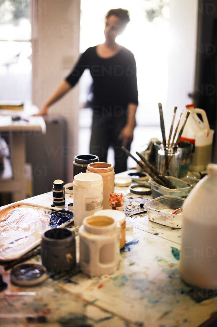 Paint bottles and containers on messy table with woman in background ...