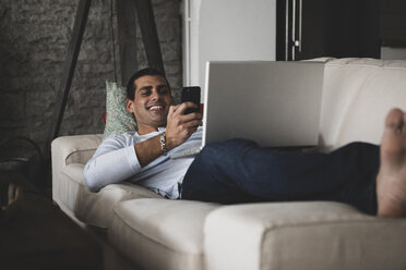 happy-young-man-lying-on-sofa-at-home-using-cell-phone-and-laptop-ERRF00359.jpg