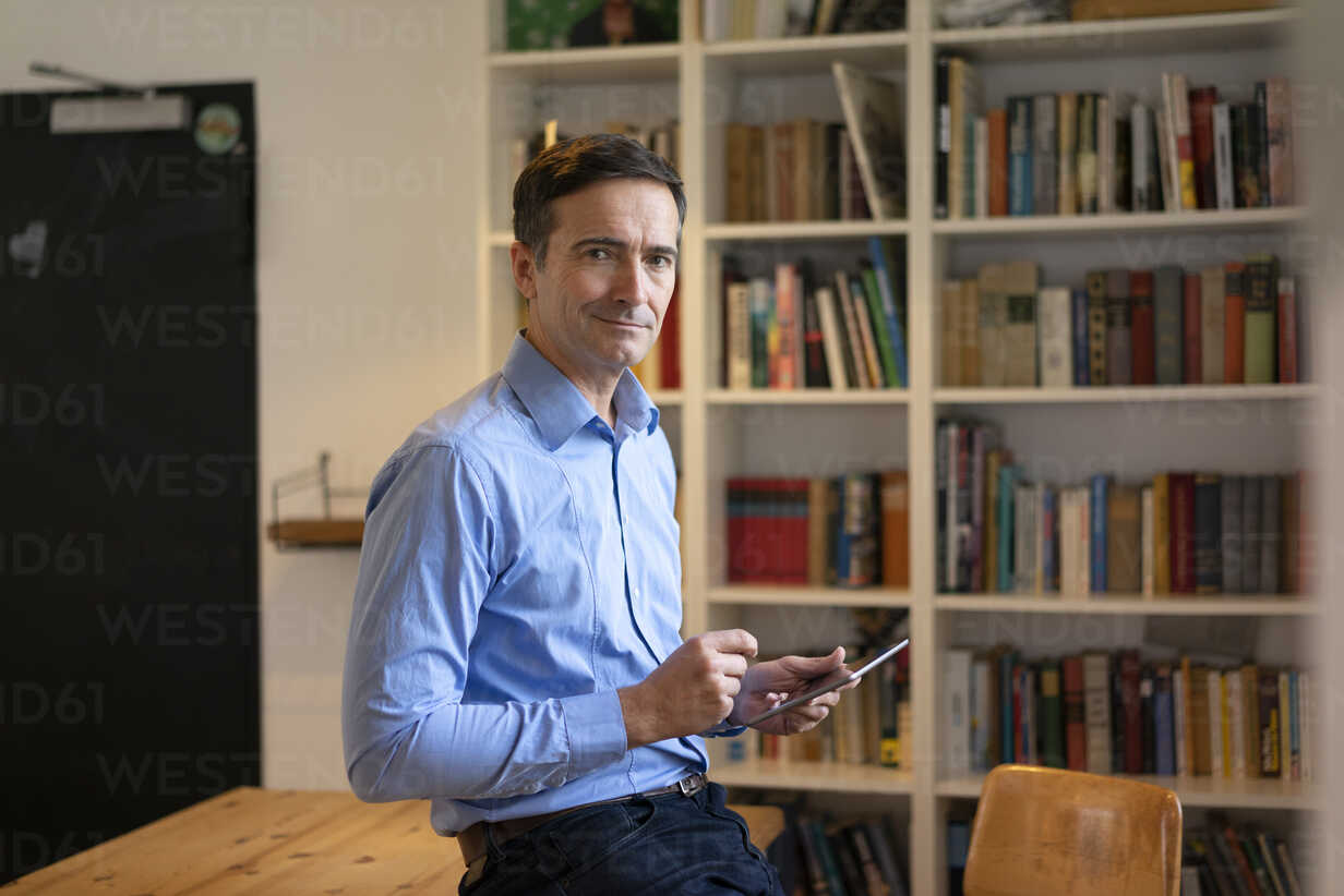 Portrait Of Confident Businessman With Tablet Leaning At Table With Bookshelf In Background Stockphoto