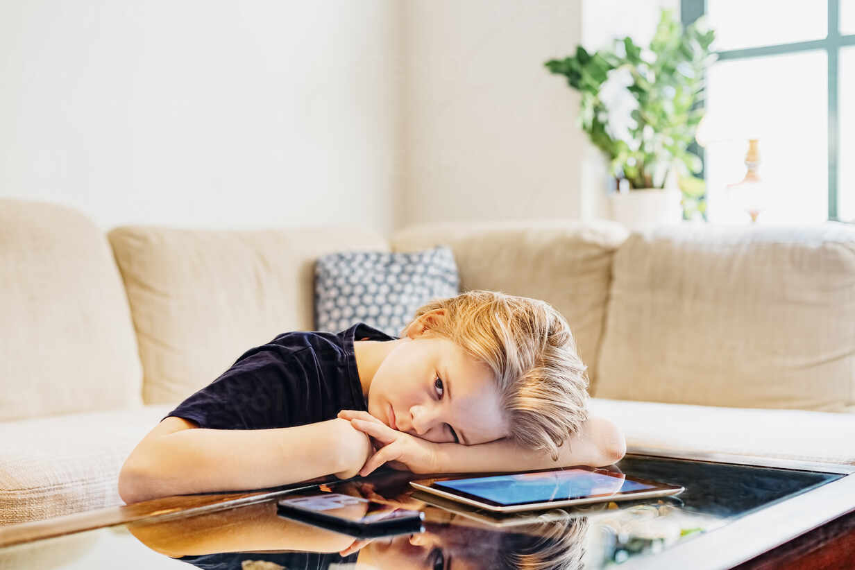 Serious Boy Lying On Coffee Table With Smartphone And Tablet Stockphoto