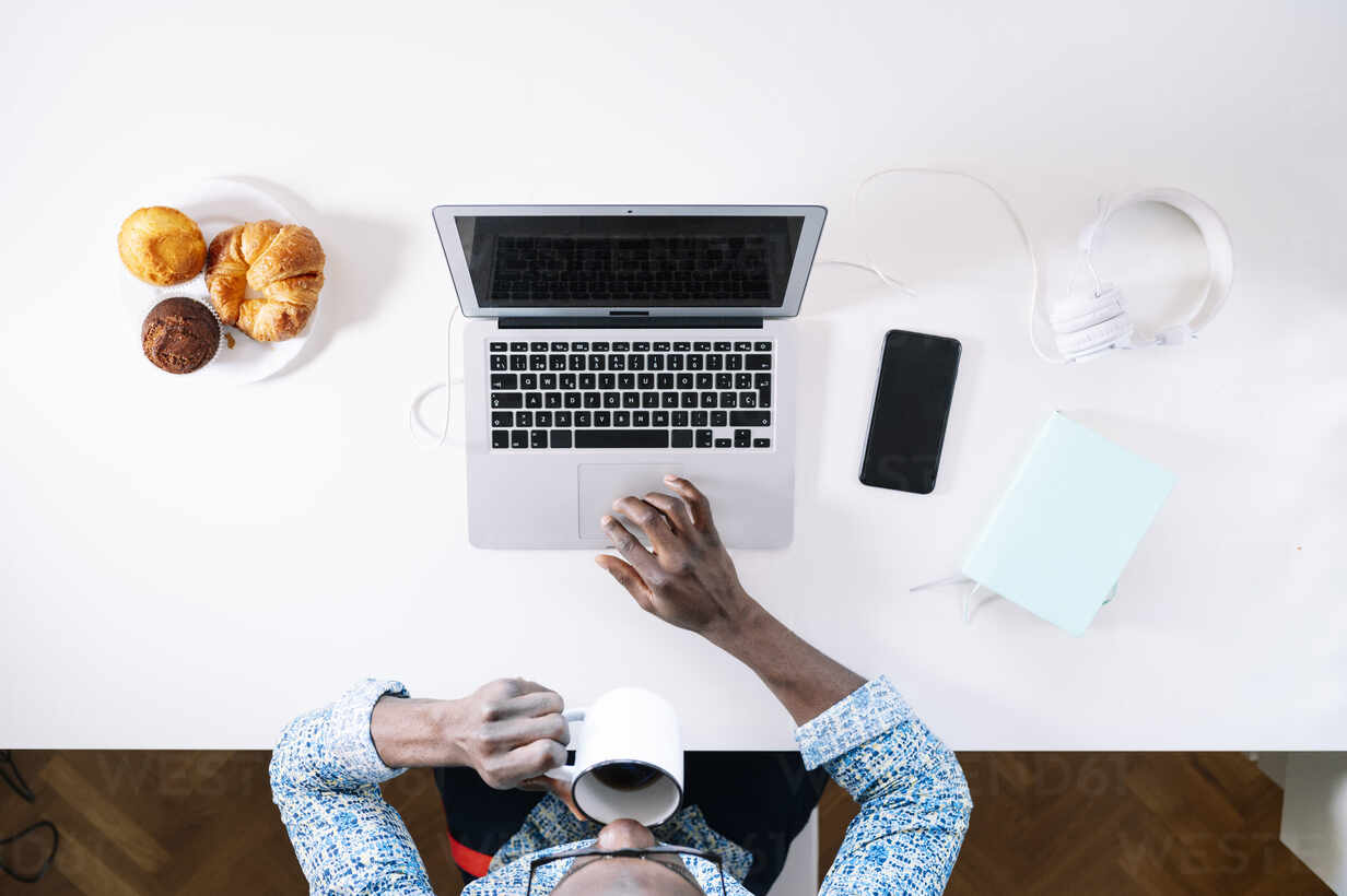 Young Man Drinking Coffee While Using Laptop At Home Office Desk Stockphoto