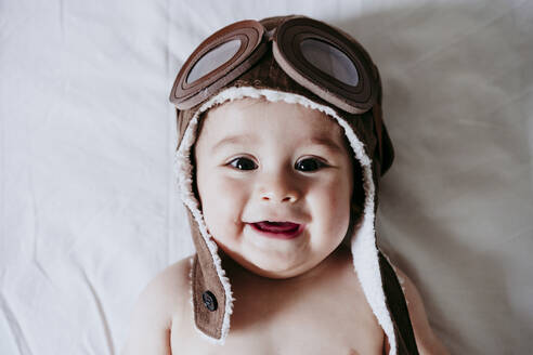 Cute Male Toddle Wearing Aviator Cap While Lying Down On Bed At Home Stockphoto